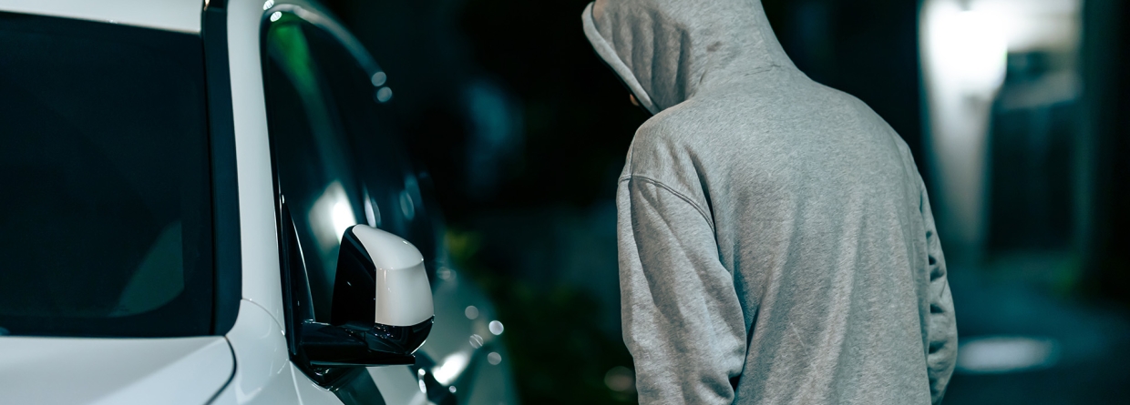 auto-inbraken; man met grijze hoodie aan en capuchon op loopt in het donker langs een witte auto en kijkt naar binnen 